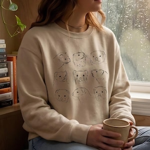 May include: Beige sweatshirt featuring a black line drawing of nine ferret faces. The crew neck sweatshirt is a neutral color and has long sleeves. The design is centered on the front of the shirt.
