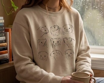 May include: Beige sweatshirt featuring a black line drawing of nine ferret faces. The crew neck sweatshirt is a neutral color and has long sleeves. The design is centered on the front of the shirt.
