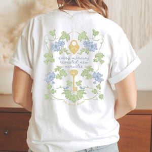 Op de afbeelding: Wit T-shirt met een bloemenpatroon en de tekst "every morning revealed new miracles" en "The Secret Garden".