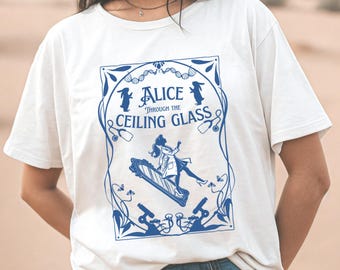 May include: White t-shirt with a blue graphic design featuring a woman in a dress holding a mirror and the text "Alice Through the Ceiling Glass".