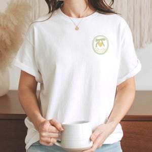 May include: A white t-shirt with a green circle design featuring a yellow heart with a green bow and the text "You're my happy place".