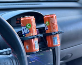 May include: Two orange soda cans in a black car cup holder. The holder is attached to the air vent in the car. The cans are labeled "C" and "NTA".