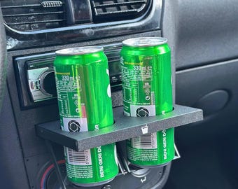 May include: Two green aluminum cans with black labels and white text reading "330 ml" are held in a black plastic car cup holder. The holder is attached to the dashboard of a car.