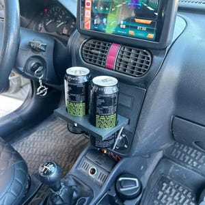 以下が含まれることがあります： 車内のカップホルダーに2つの黒と緑の飲料缶があります。車のダッシュボードには、ナビゲーション画面を表示するデジタルディスプレイがあります。インテリアはグレーとブラックで、シフトレバーとステアリングホイールが見えます。