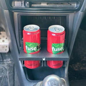May include: Two red cans of Fusetea Watermelon Flavoured Iced Tea in a black car cup holder. The text on the cans reads "Fusetea Ice Tea" and "Karpuz Aromali Icecek".