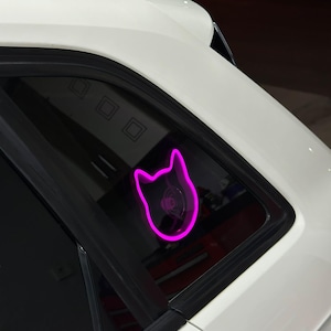 Può includere: Un adesivo per auto con la testa di un gatto rosa al neon.