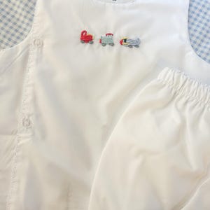 May include: White baby outfit with embroidered train, plane, and car designs. The outfit includes a sleeveless top with button closures and matching bloomers. The top has a scalloped neckline and the text "Southern Proper Blanks 12M".