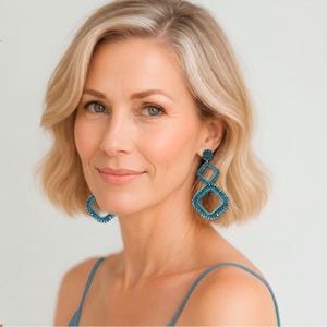 Può includere: Una donna bionda indossa orecchini geometrici turchesi. Gli orecchini hanno una forma quadrata e a diamante, con una texture di perline. Indossa un top blu con spalline sottili.