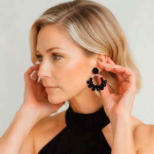 Puede incluir: Pendientes de aro con cuentas negras con un anillo de metal dorado y una parte trasera de tela negra. Los pendientes tienen un diseño circular con múltiples cuentas negras. La mujer de la imagen lleva un top negro.