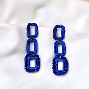 May include: A pair of handmade blue earrings. Each earring features three stacked open rectangles, with the bottom rectangle surrounded by small blue beads. The earrings are made of a woven material and hang from a small loop.