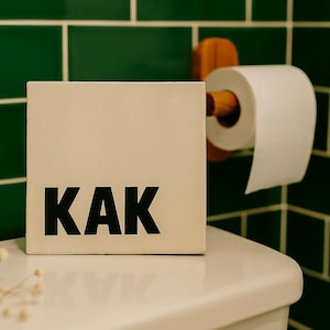 Puede incluir: Una caja blanca con la palabra "KAK" en letras negras en negrita está sobre un inodoro blanco. Un rollo de papel higiénico blanco cuelga de un soporte de madera, sobre un fondo de azulejos verdes.