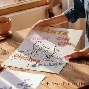 May include: A printable wall art design featuring a group of hands raising wine glasses in a toast. The words "Sant&eacute;", "Prost", "Cin Cin", "Cheers", and "Salud" are written in different colours around the glasses.