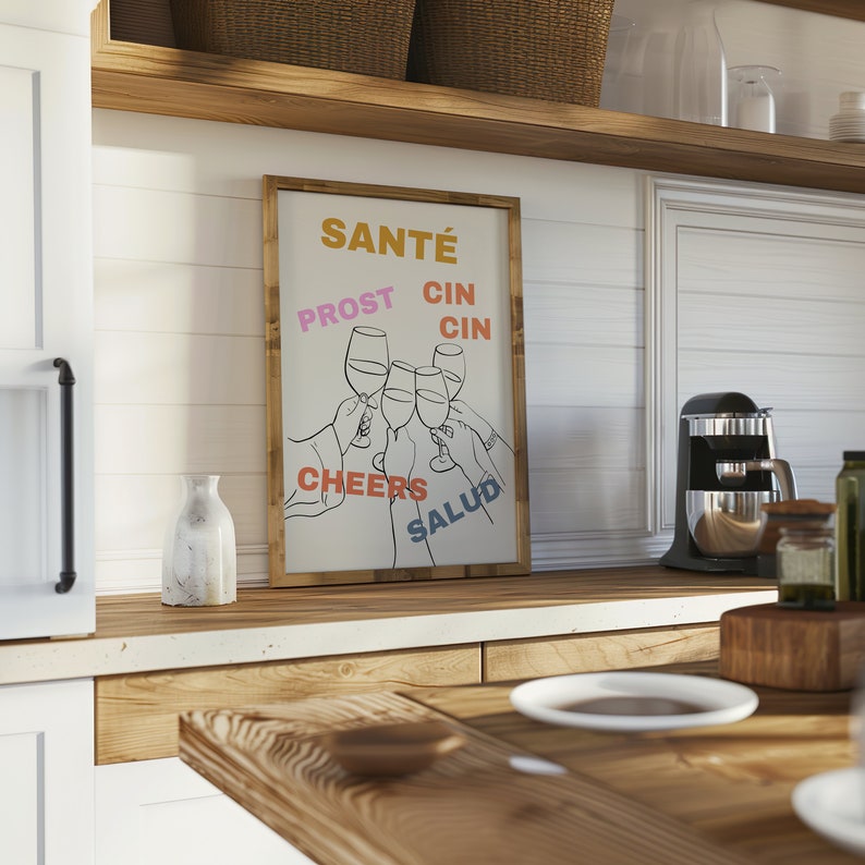 May include: A white framed print with a black line drawing of raised wine glasses. The text "Sant&eacute;", "Prost", "Cin Cin", "Cheers", and "Salud" are written in different colours around the glasses.