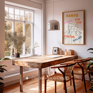 May include: A wooden table with two chairs and a framed print with the words "Sant&eacute;", "Prost", "Cin Cin", "Cheers", and "Salud" in different colours. The print features a line drawing of four hands holding up glasses of wine.
