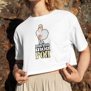 Puede incluir: Camiseta blanca recortada con un gráfico de Marilyn Monroe patinando sobre ruedas y el texto "marilyn mon roll" en amarillo.