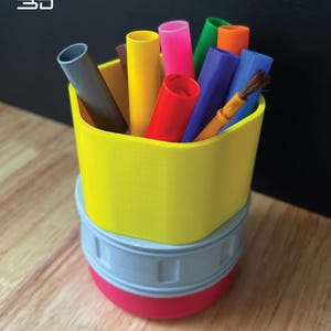 Puede incluir: Un portalápices amarillo impreso en 3D con una base gris y roja, lleno de rotuladores de colores y un pincel. El portalápices tiene un diseño curvo único. La imagen también incluye un logotipo de impresión 3D.