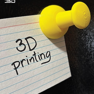 以下が含まれることがあります： 鮮やかな黄色の3Dプリントされた画鋲が、罫線入りのインデックスカードに留められています。カードには黒インクで「3D Printing」と書かれています。背景は暗く、質感のある表面です。