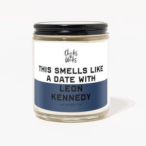 May include: A glass candle jar with a black lid and a blue label. The label says "This smells like a date with Leon Kennedy" and "Chicks & Wicks" with a heart and arrow logo. The candle weighs 7 oz.