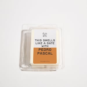 May include: A white wax melt in a clear plastic container with a label that reads "This smells like a date with Pedro Pascal". The label is orange and has the text "Chicks & Wicks" in a smaller font at the top.