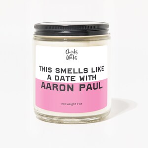 Puede incluir: Un tarro de vidrio para velas con tapa negra y una etiqueta rosa que dice "This smells like a date with Aaron Paul." La etiqueta tambi&eacute;n incluye el texto "Chicks & Wicks" y "net weight 7 oz."