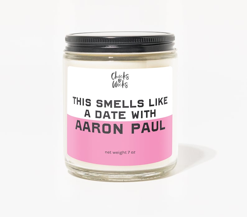 Puede incluir: Un tarro de vidrio para velas con tapa negra y una etiqueta rosa que dice "This smells like a date with Aaron Paul." La etiqueta tambi&eacute;n incluye el texto "Chicks & Wicks" y "net weight 7 oz."