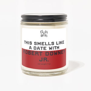 May include: A clear glass jar candle with a black lid and a white label. The label reads "THIS SMELLS LIKE A DATE WITH ROBERT DOWNEY JR." in black and red text. The candle weighs 198 grams.
