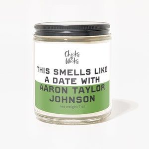 K&ouml;nnte beinhalten: Ein gr&uuml;nes und wei&szlig;es Kerzengef&auml;&szlig; mit schwarzem Deckel. Das Etikett lautet "Chicks Wicks" und "This smells like a date with Aaron Taylor Johnson" mit einem Nettogewicht von 7 oz.