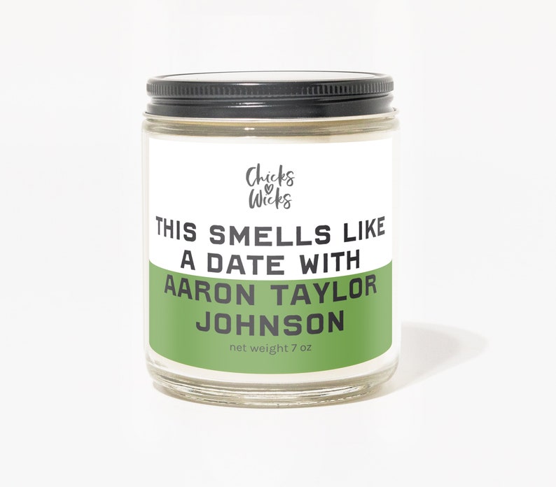 K&ouml;nnte beinhalten: Ein gr&uuml;nes und wei&szlig;es Kerzengef&auml;&szlig; mit schwarzem Deckel. Das Etikett lautet "Chicks Wicks" und "This smells like a date with Aaron Taylor Johnson" mit einem Nettogewicht von 7 oz.