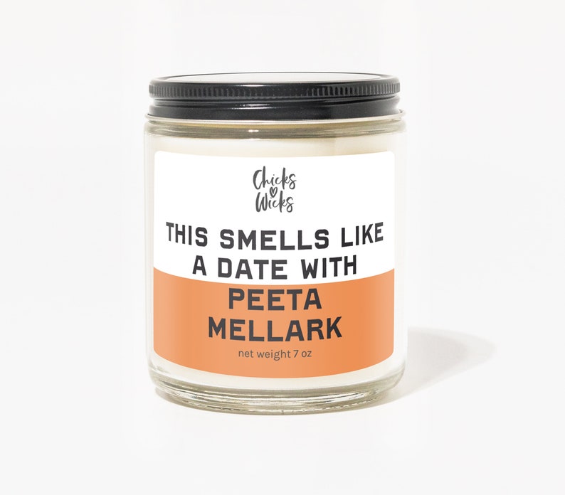 K&ouml;nnte beinhalten: Ein wei&szlig;es Kerzengef&auml;&szlig; mit schwarzem Deckel. Das Etikett lautet "Chicks & Wicks" und "This smells like a date with Peeta Mellark". Das Gef&auml;&szlig; ist 7 oz.