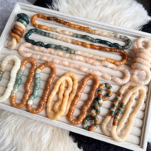 May include: A collection of beaded necklaces and bracelets in various colors, including cream, brown, green, and orange. The jewelry is displayed in a white tray with a soft, textured background.