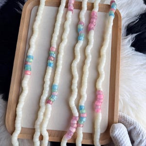 May include: Four off-white beaded necklaces with sections of pink, blue, and iridescent beads. The necklaces are displayed on a wooden tray, with a white and black patterned background.