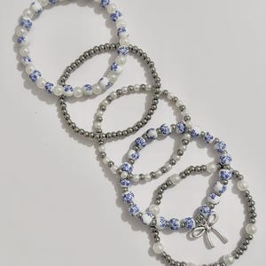 May include: Five beaded bracelets. The bracelets feature a mix of white faux pearls, silver-colored beads, and white beads with blue floral patterns. One bracelet has a silver-colored bow charm.