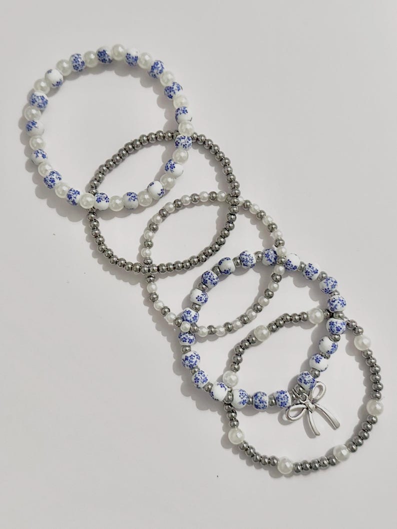May include: Five beaded bracelets. The bracelets feature a mix of white faux pearls, silver-colored beads, and white beads with blue floral patterns. One bracelet has a silver-colored bow charm.