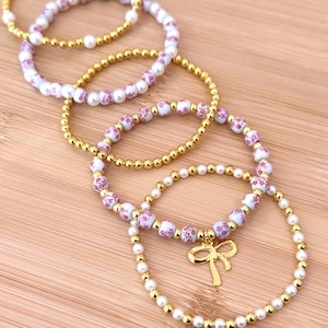 May include: A collection of beaded bracelets in gold and white. Some bracelets feature small, round gold beads, while others have white beads with a pink floral pattern. One bracelet has a gold bow charm.
