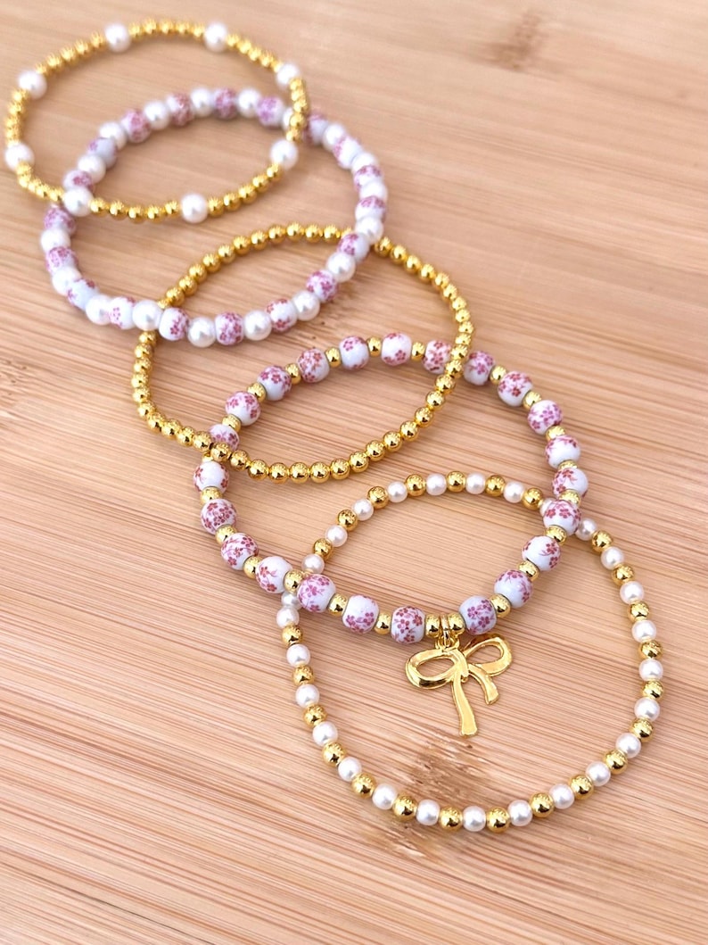 May include: A collection of beaded bracelets in gold and white. Some bracelets feature small, round gold beads, while others have white beads with a pink floral pattern. One bracelet has a gold bow charm.
