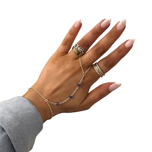 Può includere: Una mano ornata di anelli d'oro e una delicata catena per la mano dorata. Un anello presenta la scritta "DIOR". La catena per la mano, collegata a un braccialetto, è impreziosita da piccole perle blu. Le unghie sono dipinte di rosa chiaro.