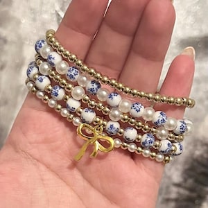 May include: A stack of beaded bracelets in gold, white, and blue. The bracelets feature gold beads, white pearls, and white beads with blue floral patterns. A gold bow charm hangs from one bracelet.