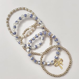 May include: Several beaded bracelets in a variety of styles. Some bracelets feature gold-colored beads, while others have white beads with blue floral patterns. One bracelet has a gold bow charm.