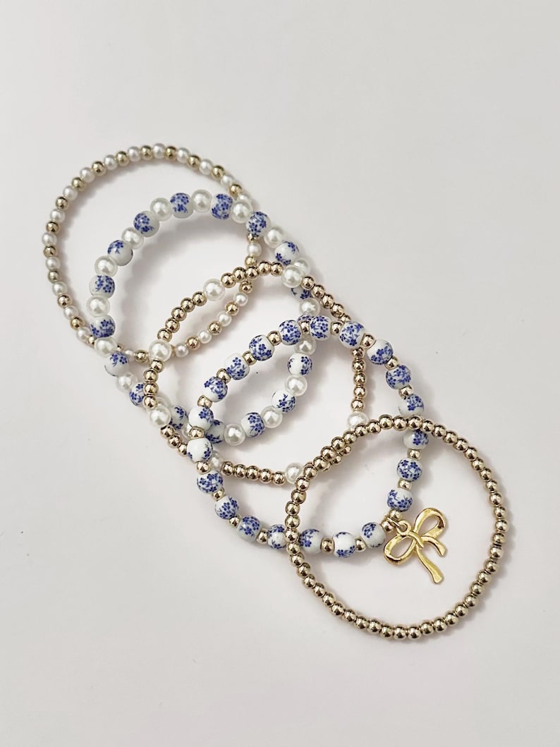 May include: Several beaded bracelets in a variety of styles. Some bracelets feature gold-colored beads, while others have white beads with blue floral patterns. One bracelet has a gold bow charm.