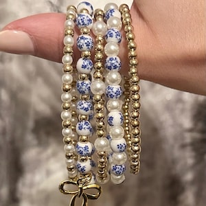 May include: A selection of beaded bracelets in gold, white, and blue. The bracelets feature gold beads, white faux pearls, and blue and white floral patterned beads. A gold bow charm hangs from one bracelet. The bracelets are stacked together.