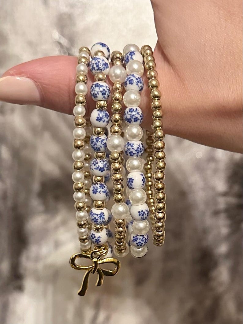 May include: A collection of beaded bracelets in gold, white, and blue. The bracelets feature gold beads, white faux pearls, and blue and white floral patterned beads. A gold bow charm hangs from one bracelet. The bracelets are stacked together.