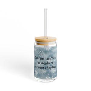Peut inclure: Un verre transparent avec un couvercle en bambou et une paille transparente. Le verre présente un fond bleu nuageux avec des illustrations de flocons de neige blancs et le texte noir : Current location: somewhere between chapters.