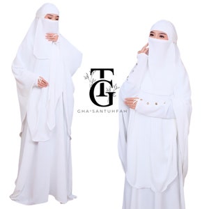 Peut inclure: Abaya blanche avec niqab et manches longues. L'abaya a un design fluide et est faite d'un tissu doux et léger. Le niqab est attaché à l'abaya et couvre le visage. Les manches ont un détail de bouton au poignet. Le texte "GHA'ISANTUHFAH" est visible sur l'image.