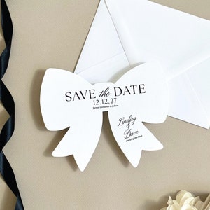 May include: White save the date card shaped like a bow with the text "Save the Date" and the date "12.12.27". Below the date is the text "Formal invitation to follow". The names "Lindsey & Dave" are written in cursive with the text "are tying the knot" below.