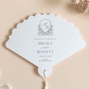 May include: A white fan-shaped wedding program card with a scalloped edge and a cream-colored tassel. The text reads: Welcome to the wedding of Mikaila and Bennett, April 30, 2028, El Chorro, Paradise Valley, Arizona, with a floral MB monogram.
