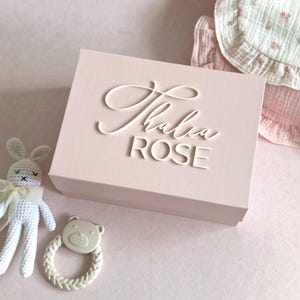 May include: A light pink gift box with the name "Thalia Rose" in white script. A white crocheted bunny and a bear-shaped rattle are next to the box. A pink bib with a floral pattern is in the background.