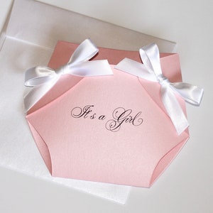 May include: Pink baby shower invitation shaped like a diaper with white satin bows and the text "It's a Girl" in black cursive.
