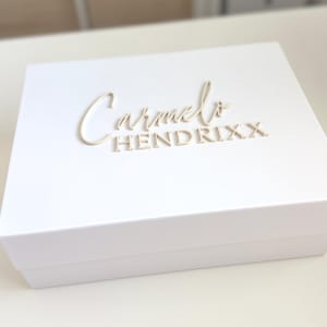 May include: A white box with the name "Carmelo Hendrixx" in a light beige, elegant font. The box is rectangular and appears to be made of a sturdy material. The background is blurred, focusing attention on the box.