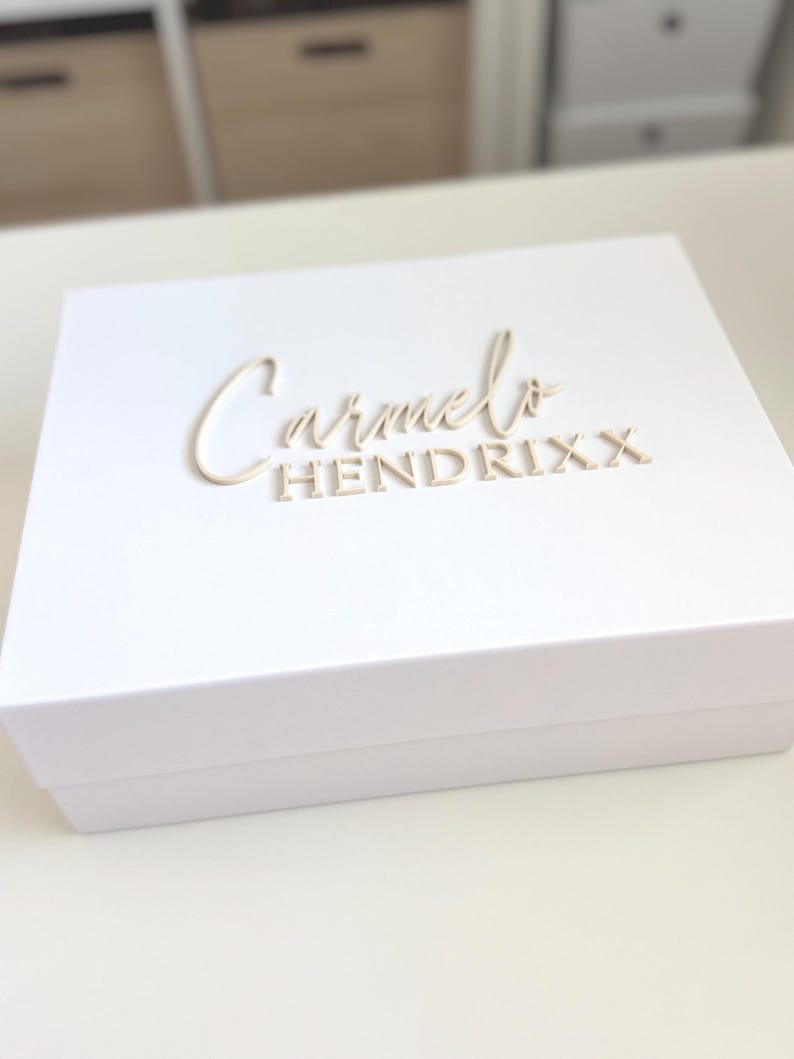 May include: A white box with the name "Carmelo Hendrixx" in a light beige, elegant font. The box is rectangular and appears to be made of a sturdy material. The background is blurred, focusing attention on the box.