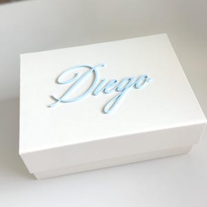 May include: A white gift box with a light blue cursive "Diego" on the lid. The box is rectangular and sits on a white surface. The text is in a delicate, flowing script.